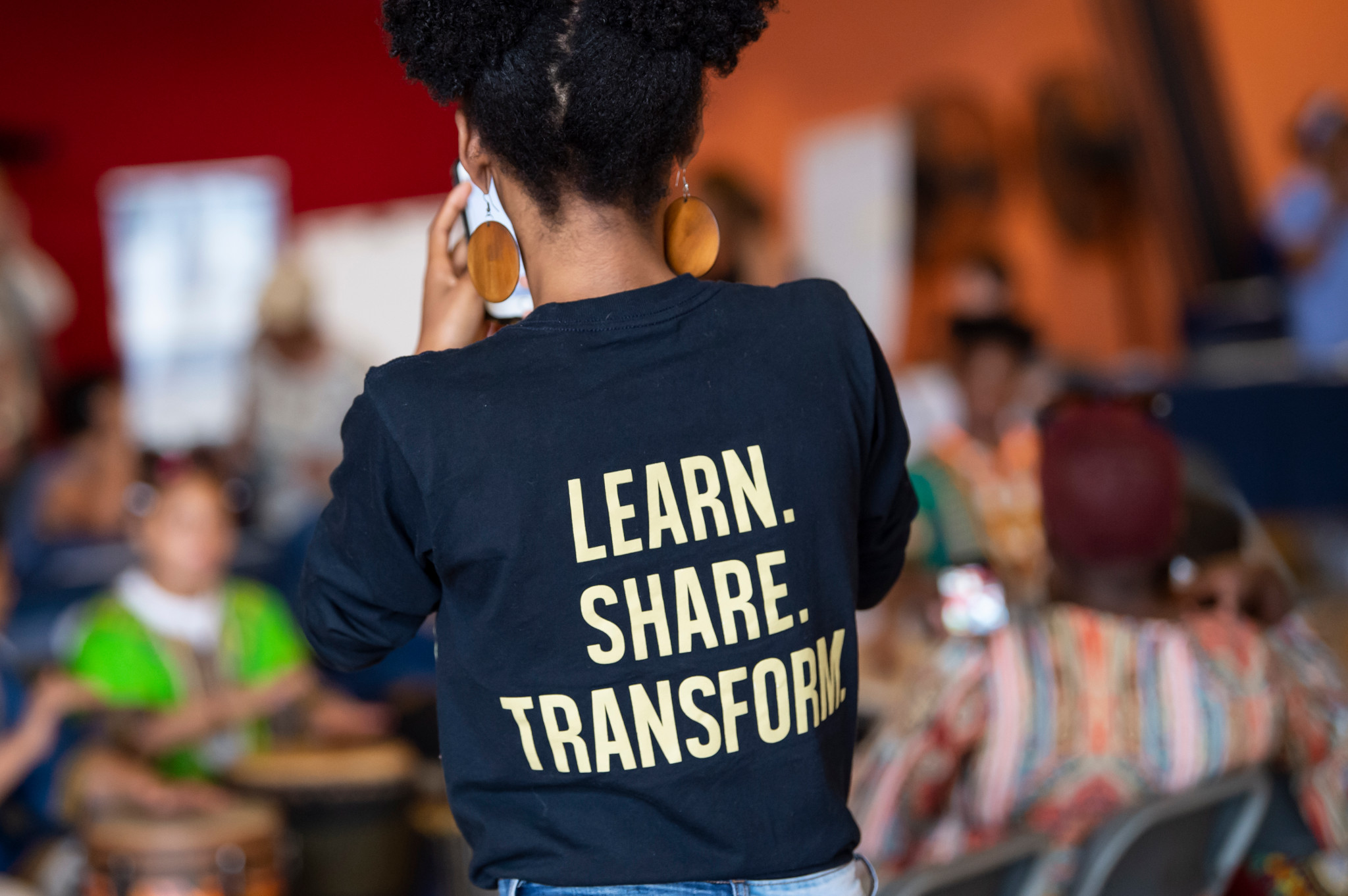 the back of a standing individual wearing a shirt with the words "LEARN. SHARE. TRANSFORM."