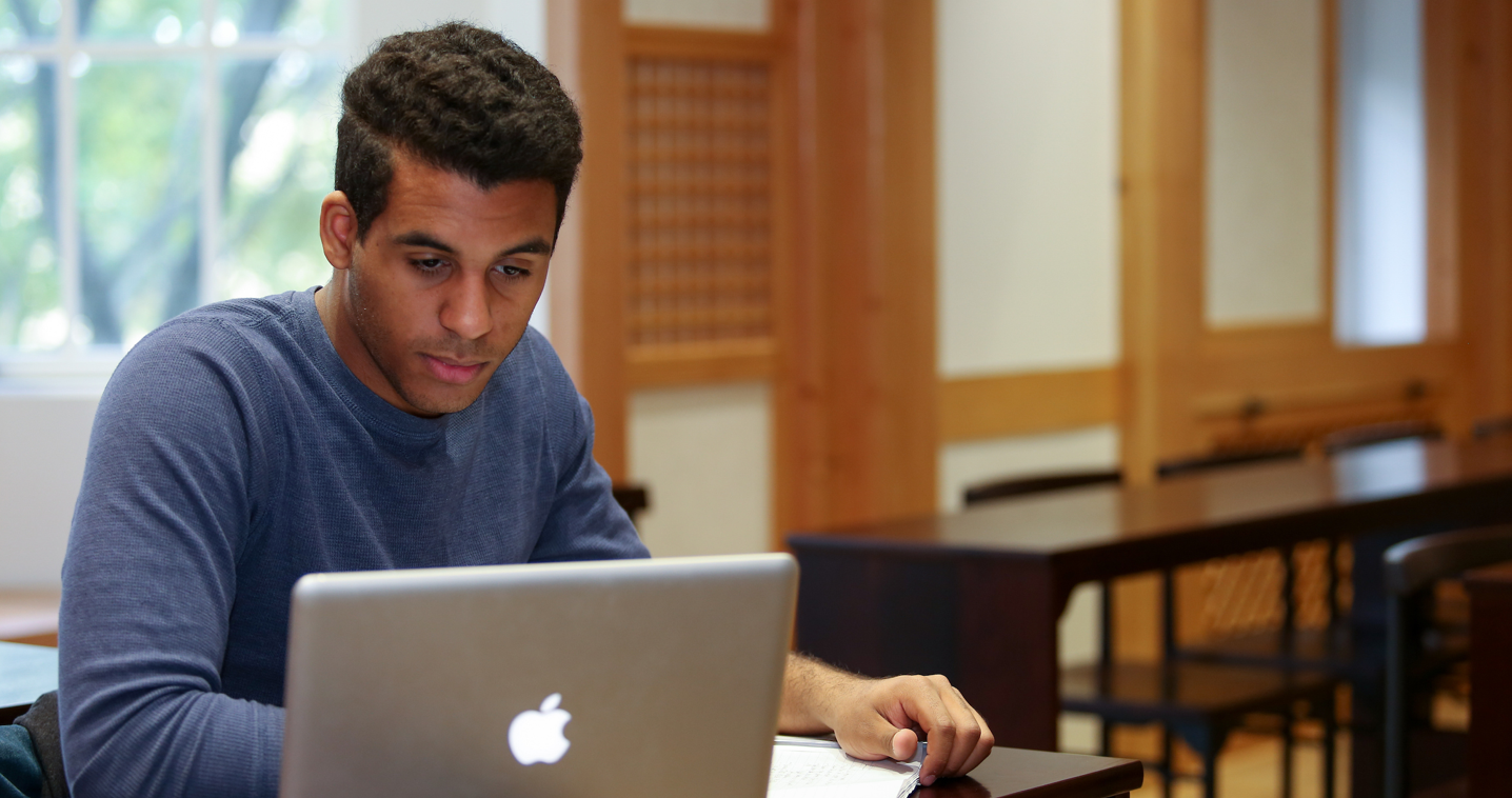 Male student at a table looking down at his laptop screen