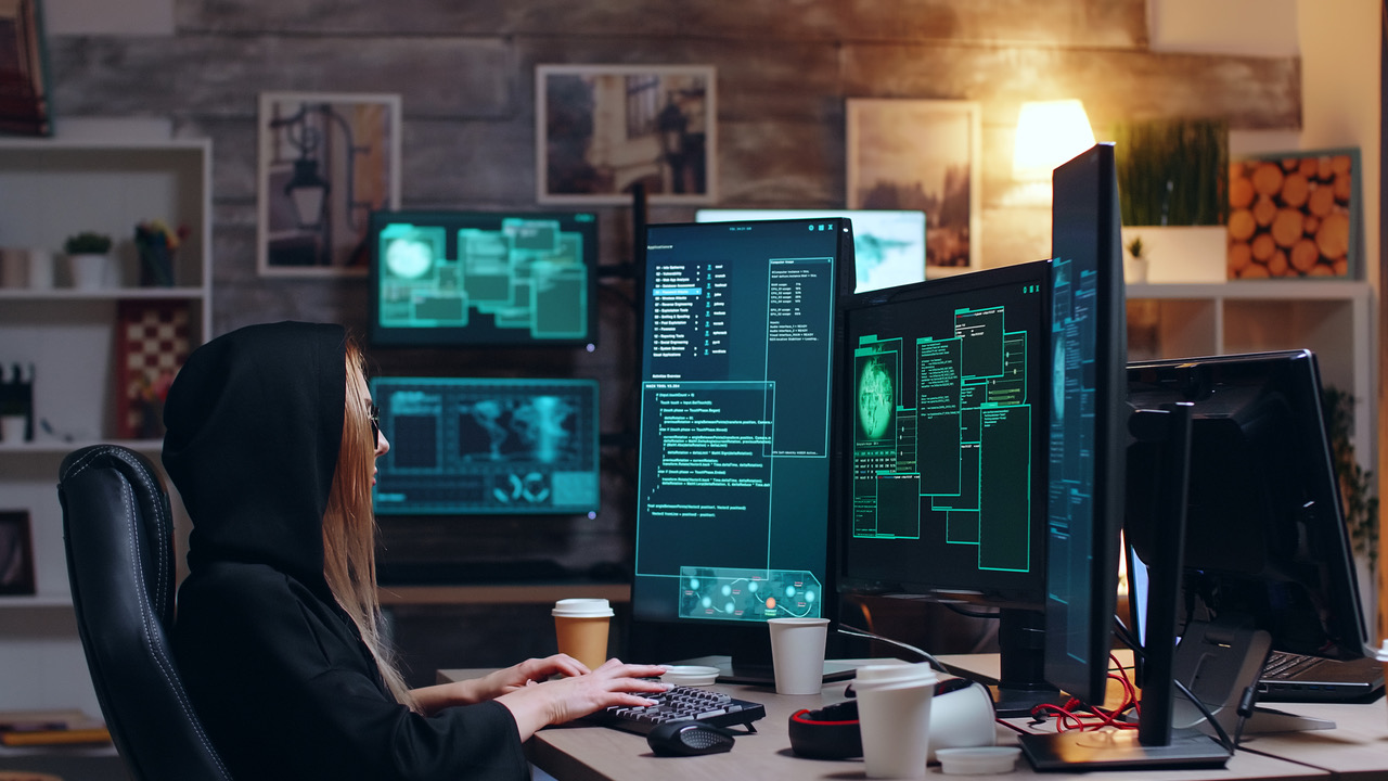 an individual with a hoodie sitting in front of 5 computer screens 