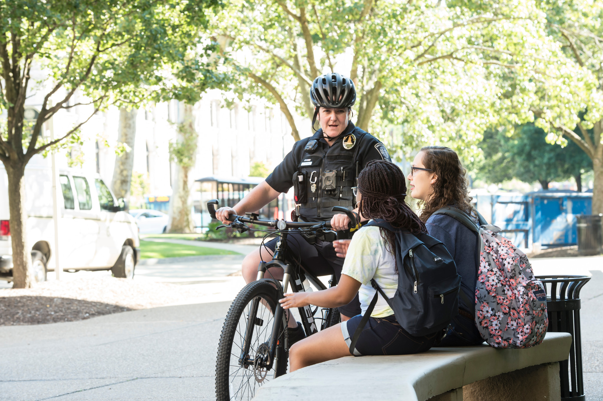 a police officer on a bicycle talking to two students sitting by a bench