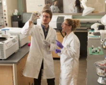 Two students wearing lab coats and gloves in lab